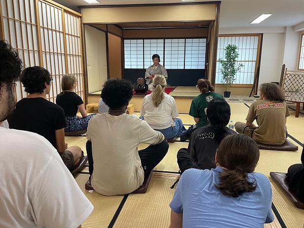 View of students sitting on floor cross legged from back of the room with tea ceremony hostess facing them